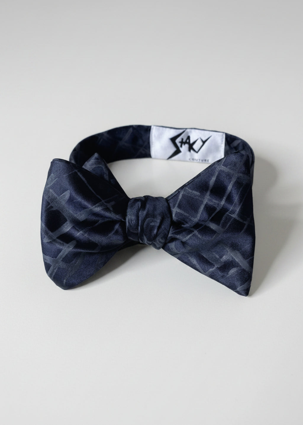 A silk hand-painted black plaid bow tie by Stacy Stewart Smith, front view showing brand label against a neutral background.