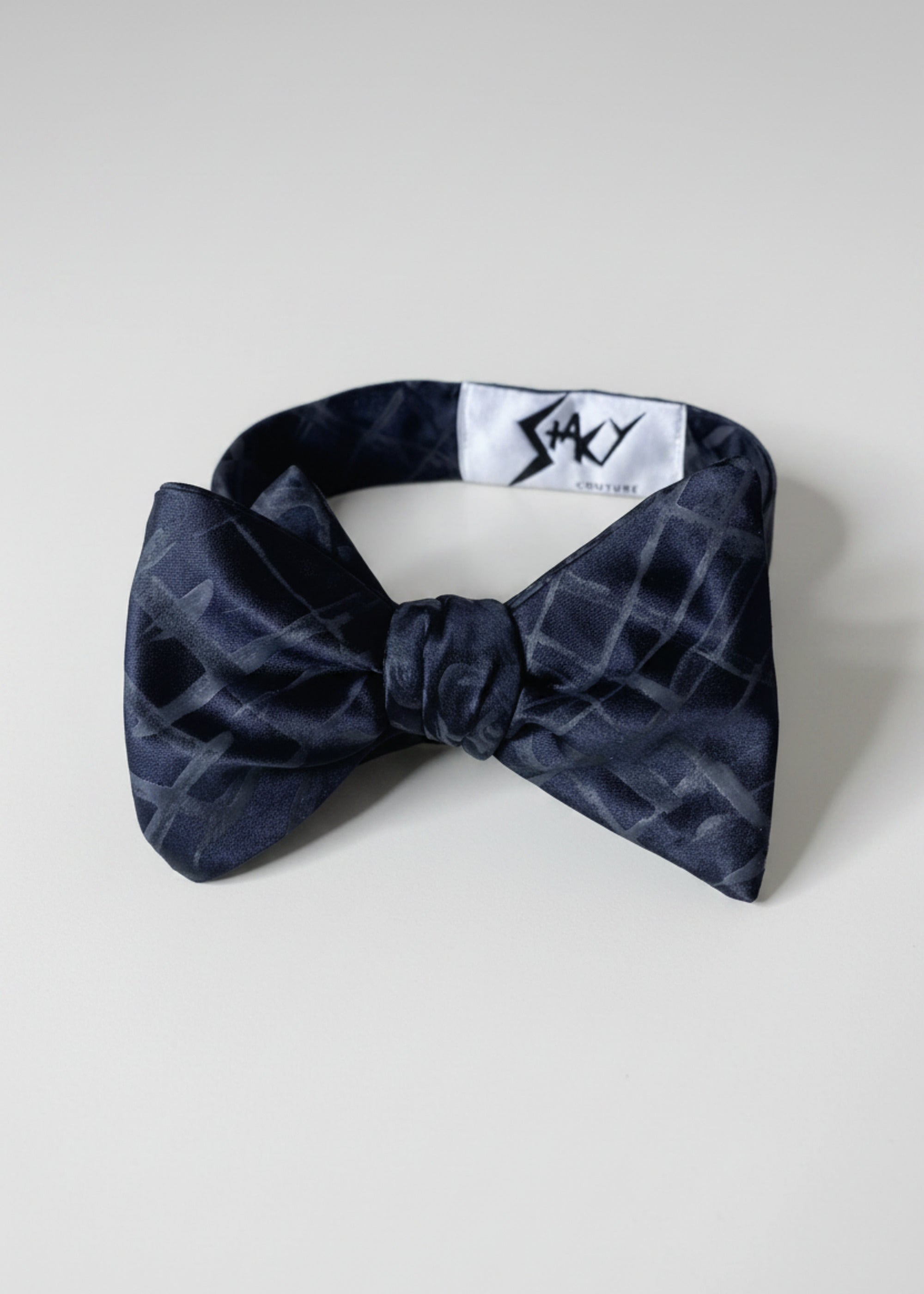 A silk hand-painted black plaid bow tie by Stacy Stewart Smith, front view showing brand label against a neutral background.