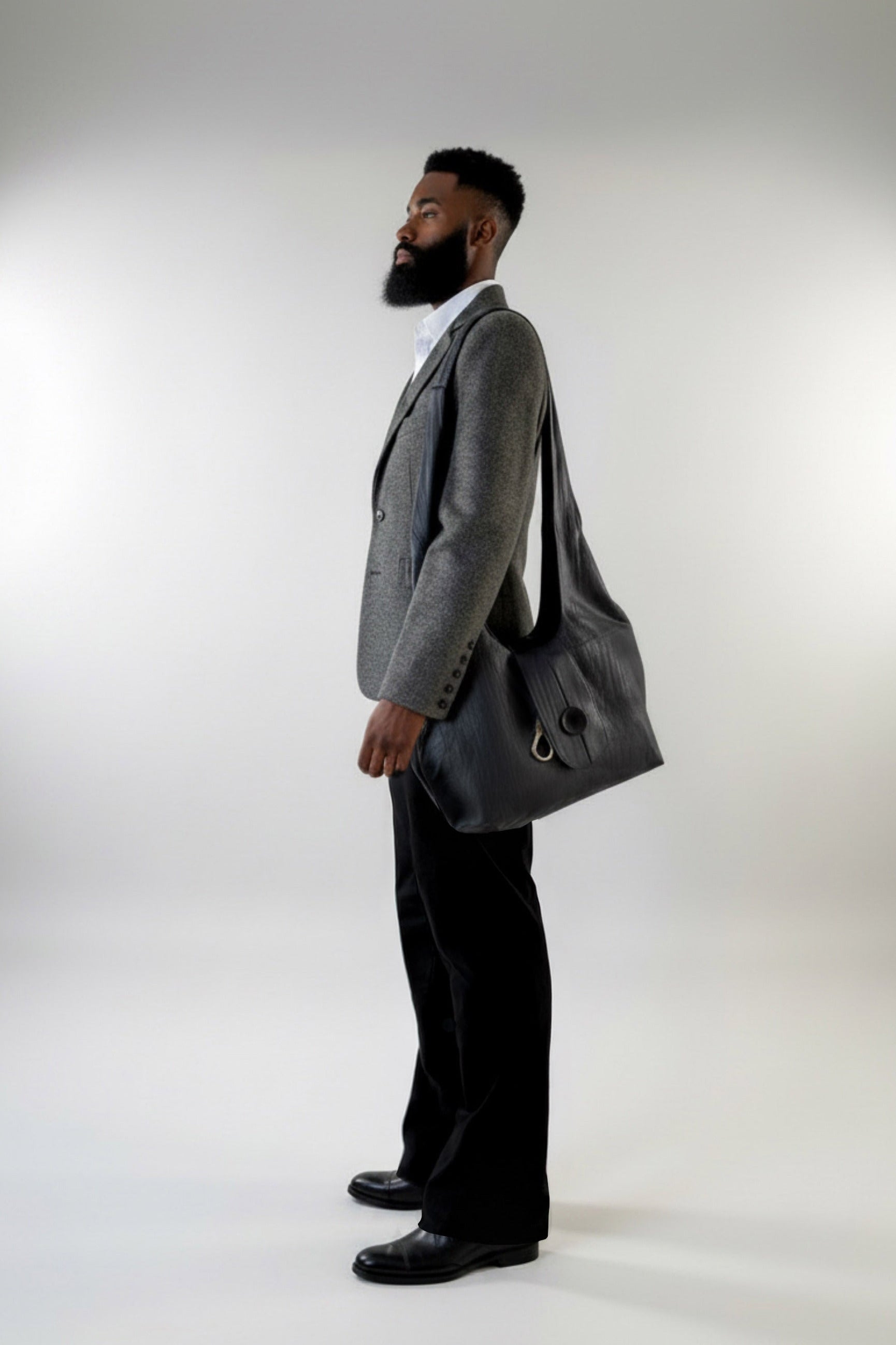 A man in a Diamond Dodger tweed blazer, white shirt, and black Windsor Pants by Stacy Stewart Smith stands in profile carrying a large black crinkled leather Euclidean Space messenger bag also, by the  menswear couture designer, against a light gray studio background.