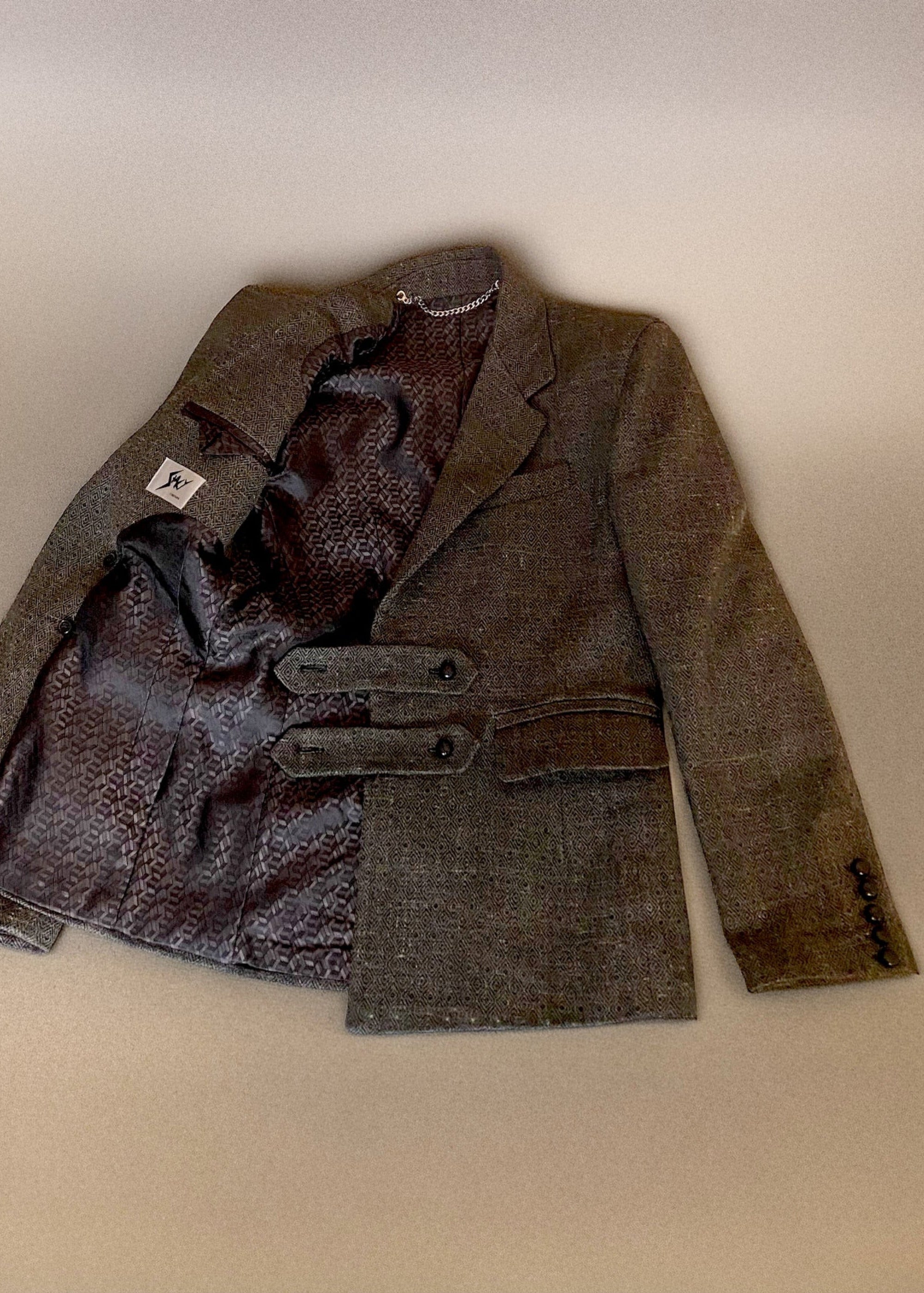 Stacy Stewart Smith Men's Highland Gentry Diamond Tweed Suit lying flat with one side of the front open revealing the box pattered acetate lining, the Stacy Couture brand label and chain hanging accessory on an Ivory shadowed paper surface. 