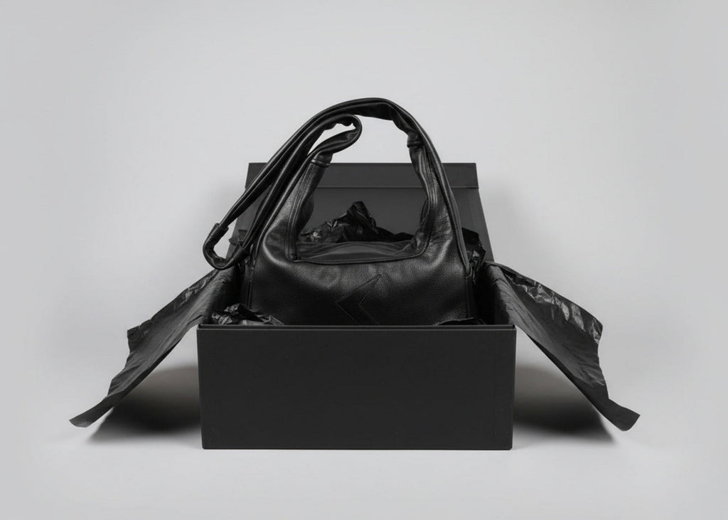 Stacy Stewart Smith Trapezoid Leather Messenger Bag in Onyx sitting in a black gift box surrounded by black tissue paper in front of a light gray backdrop.