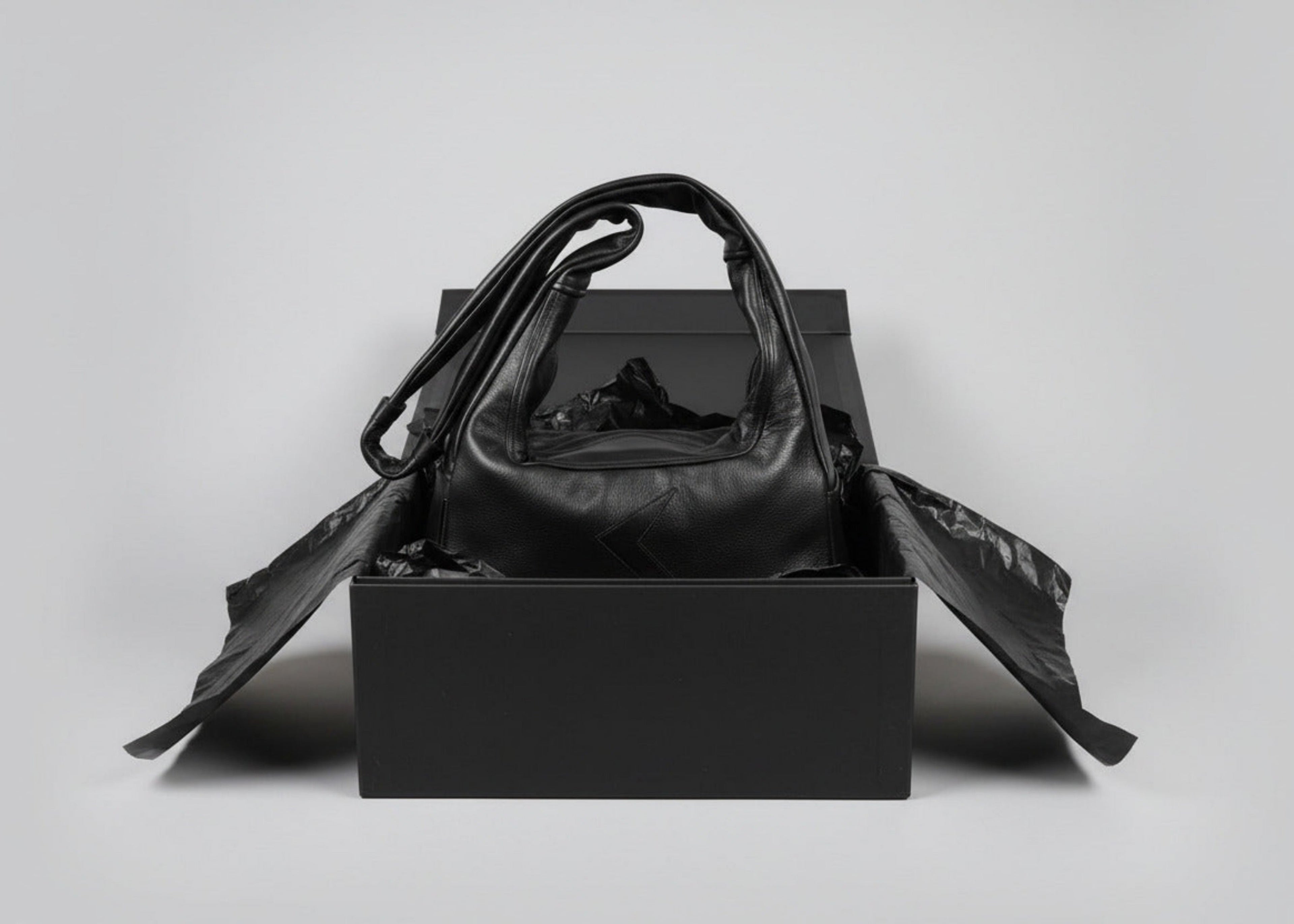 Stacy Stewart Smith Trapezoid Leather Messenger Bag in Onyx sitting in a black gift box surrounded by black tissue paper in front of a light gray backdrop.