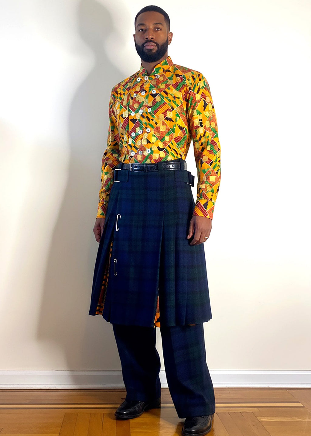 A handsome man modeling Stacy Stewart Smith's Blackwatch Plaid Harris Tweed © Reality Kilt over matching plaid pleated Windsor Pants With the Noble Kente cloth cotton shirt featuring a vibrant multicolor tribal motif standing against a white wall on a wooden floor.