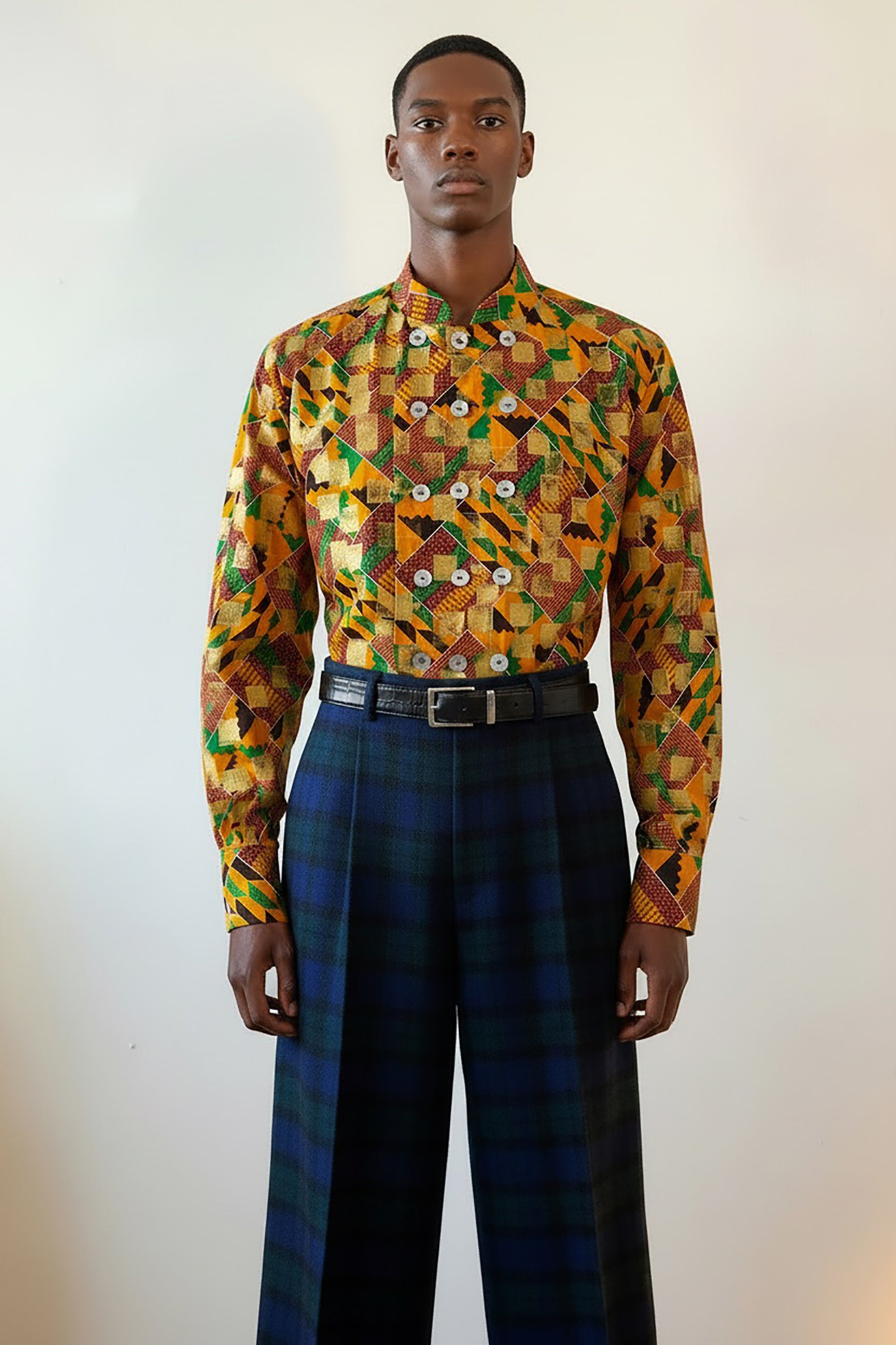 Stacy Stewart Smith's Noble Kente long-sleeve button-up premium cotton Kente cloth dress shirt with manadarin collar, long sleeve gold metallic hand pixel over-printing and real shell buttons styled with Blackwatch plaid Windsor pants on a male model torso front view against a plain wall.