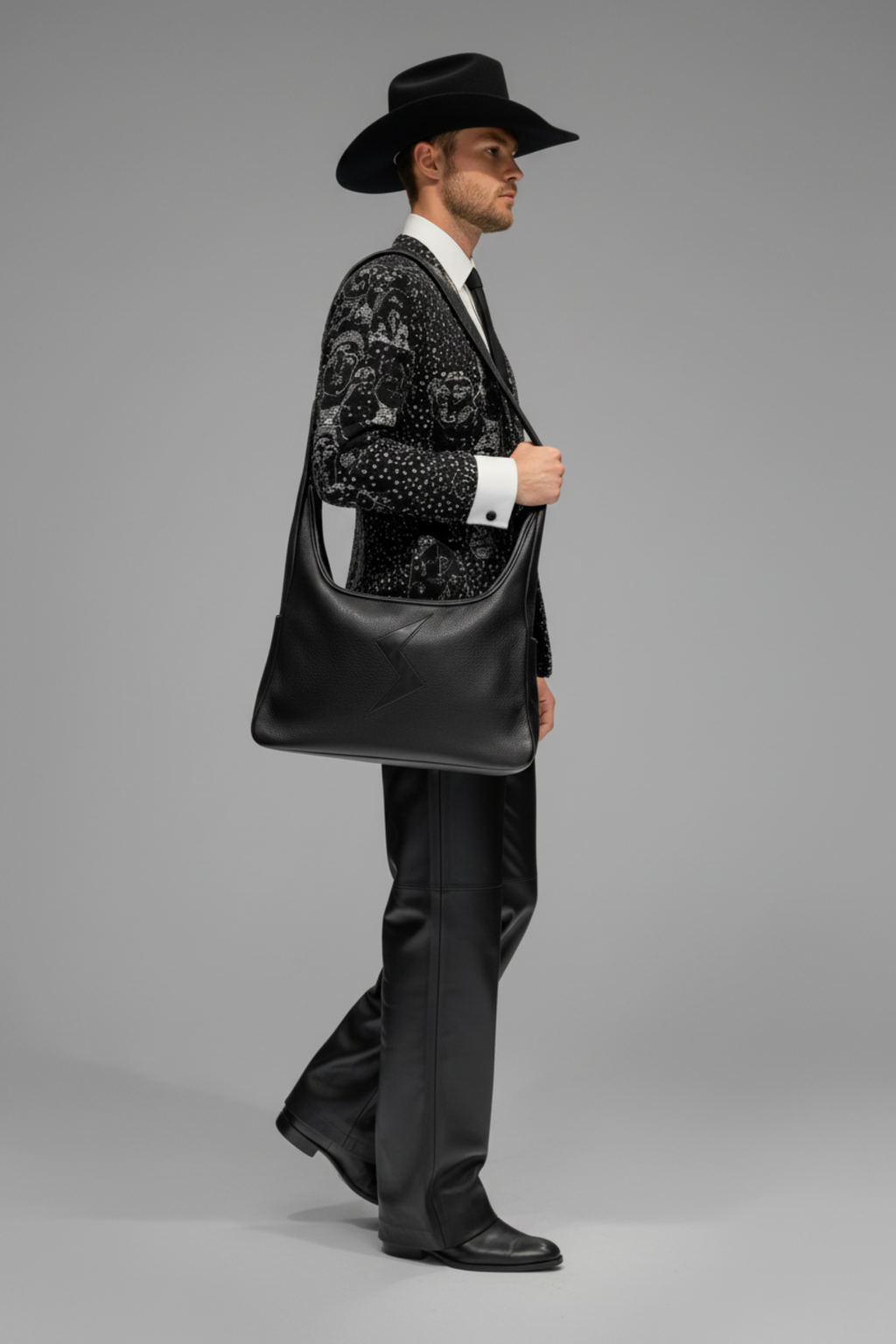 A man in a black cowboy hat, patterned black and white jacket, white shirt, and black trousers carries the Stacy Stewart Smith Black leather Trapezoid Messenger Bag against a neutral gray studio background.