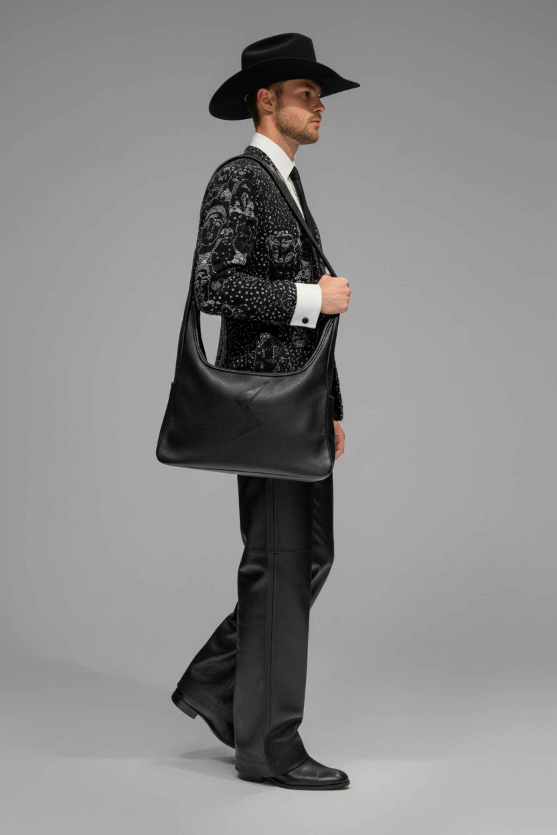 A man in a black cowboy hat, patterned black and white jacket, white shirt, and black trousers carries the Stacy Stewart Smith Black leather Trapezoid Messenger Bag against a neutral gray studio background.