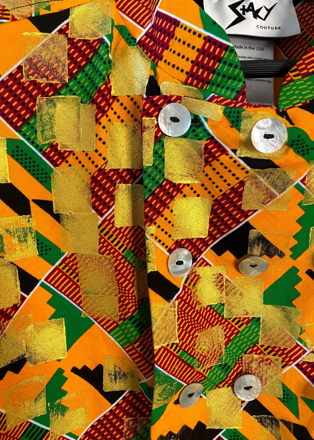 Neckline close up of Stacy Stewart Smith's Noble Kente long-sleeve button-up premium cotton Kente cloth dress shirt with manadarin collar, long sleeve gold metallic hand pixel over-printing with natural shell buttons with brand label Stacy Couture Made in the USA showing.