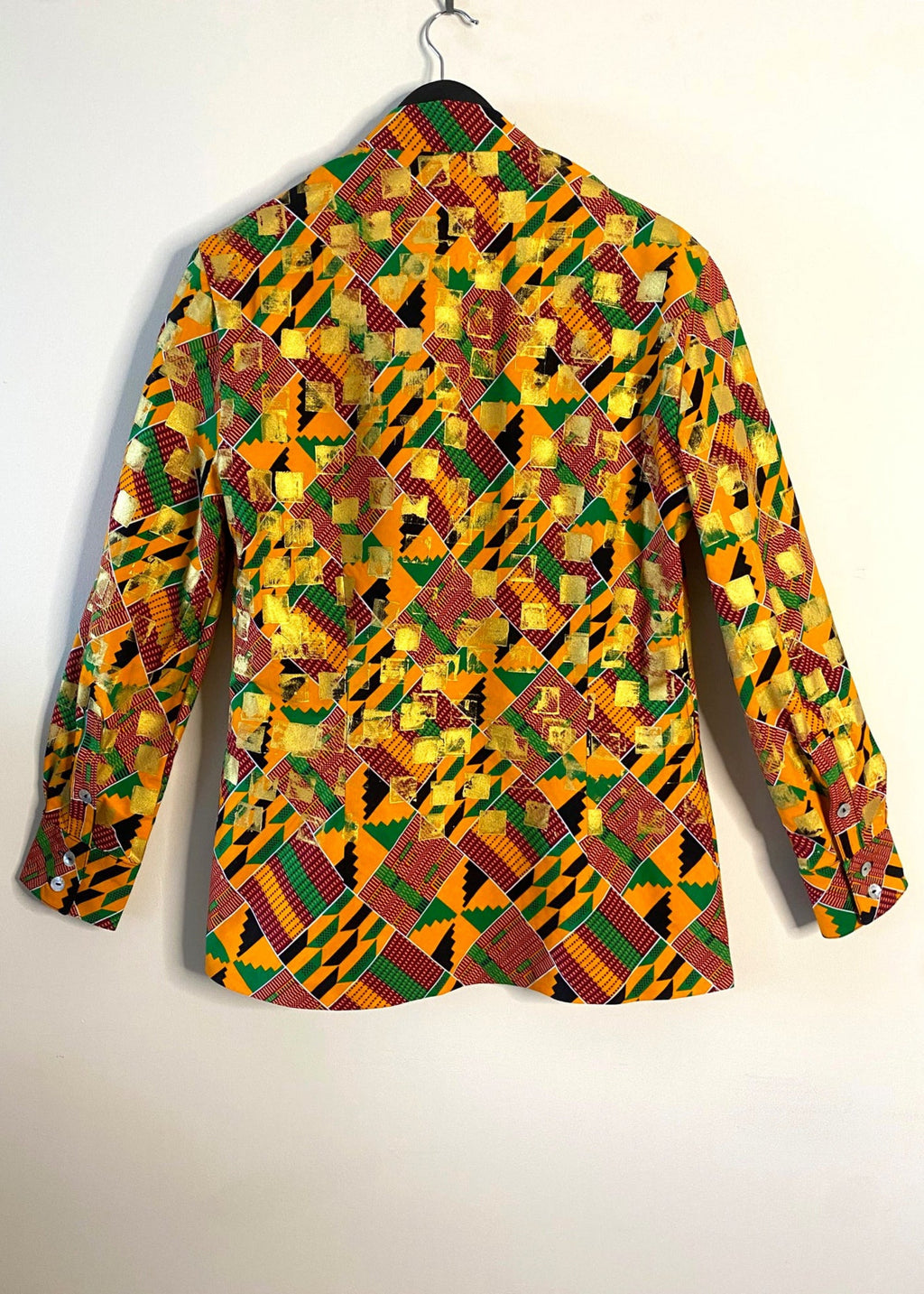 Stacy Stewart Smith's Noble Kente long-sleeve button-up premium cotton Kente cloth dress shirt with manadarin collar, long sleeve gold metallic hand pixel over-printing with natural shell buttons displayed on a hanger back view against a plain wall.