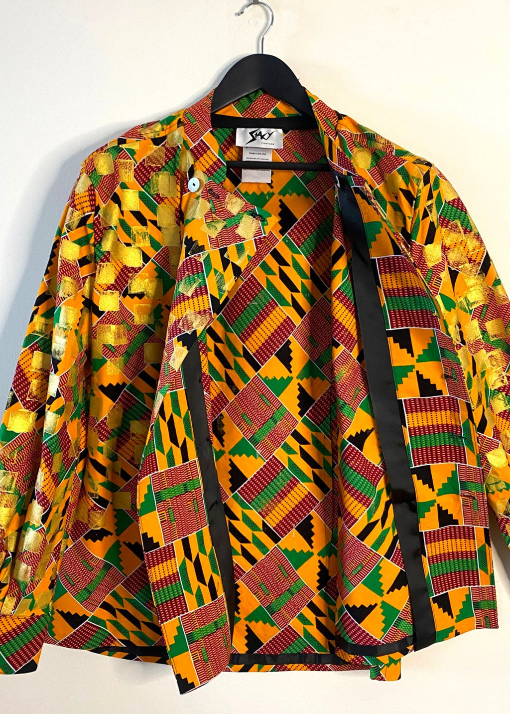 Stacy Stewart Smith's Noble Kente long-sleeve button-up premium cotton Kente cloth dress shirt with manadarin collar, long sleeve gold metallic hand pixel over-printing with natural shell buttons displayed on a hanger front open view revealingg couture construction and brand label "Stacy couture" against a plain wall.