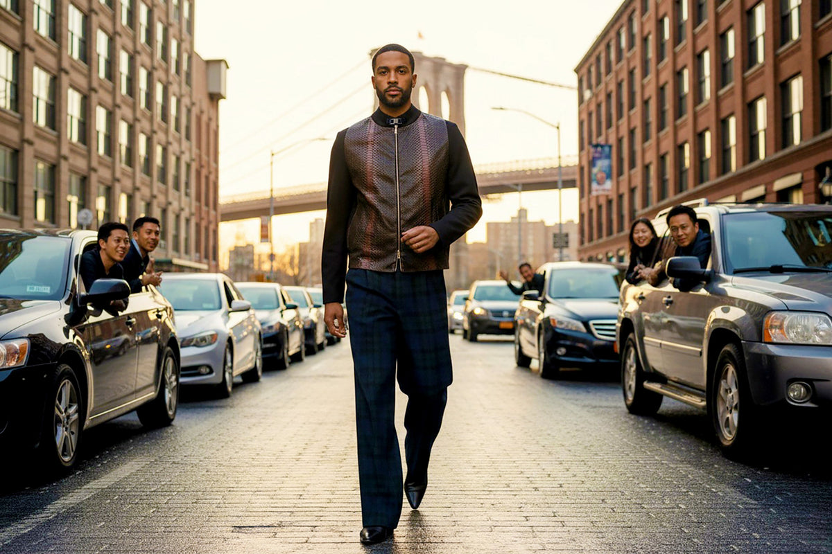 Stacy Stewart Smith's Python & Denim Bellhop Jacket with Blackwatch Plaid Harris Tweed Reality Pants on male model 6'3" in Dumbo, New York with the Brooklyn Bridge in the distance walks on a cobblestone street while people in cars near brick buildings on both sides look toward him in admiration.