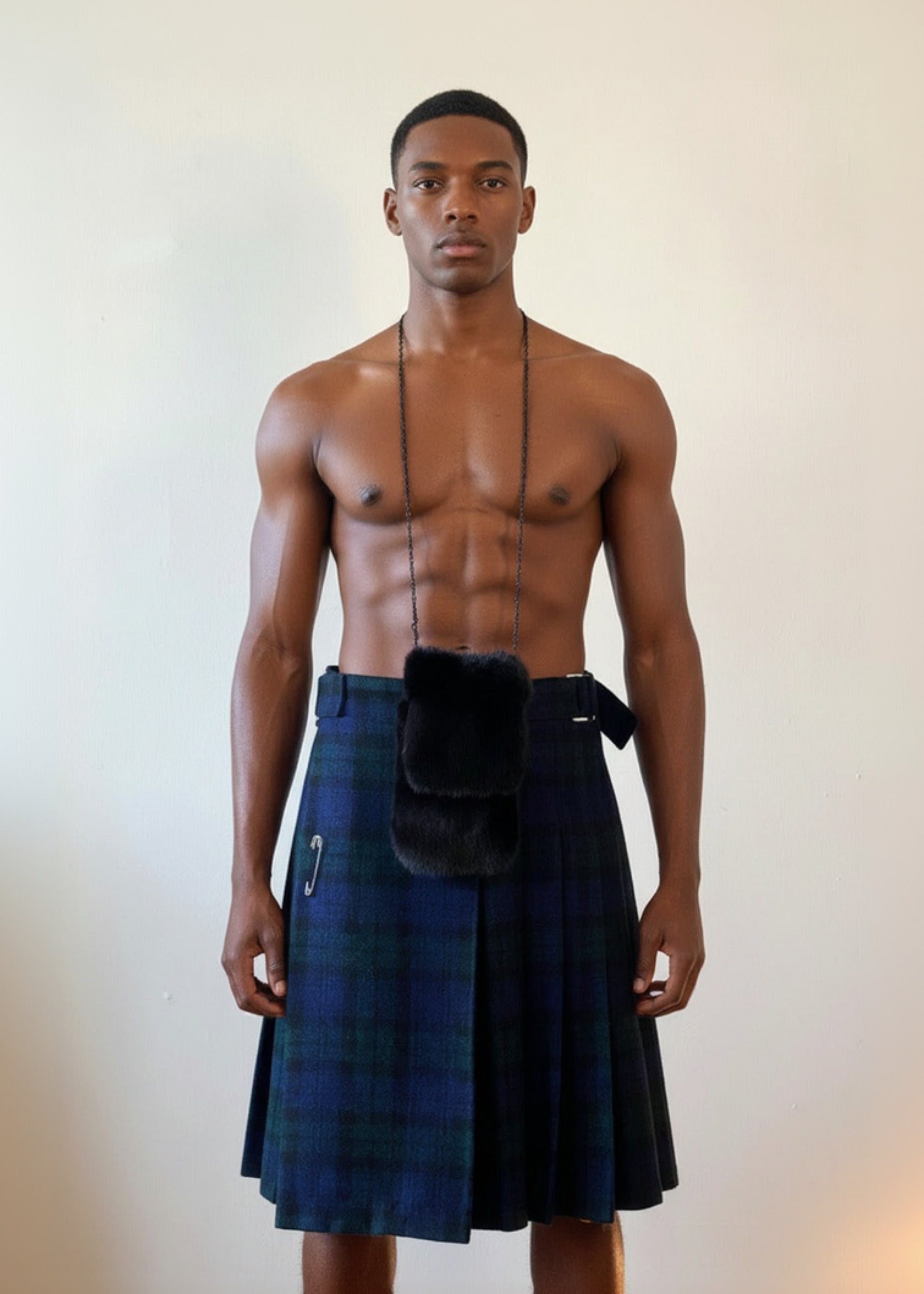 Stacy  Stewart Smith men's Harris Tweed© Blackwatch in blue and green tones Plaid reality Kilt has Kente cloth inlays, chrome side adjustable buckles and kilt pin worn with Smith's Black Gamma Mink sporran bag on shirtless male model against a plain light wall.