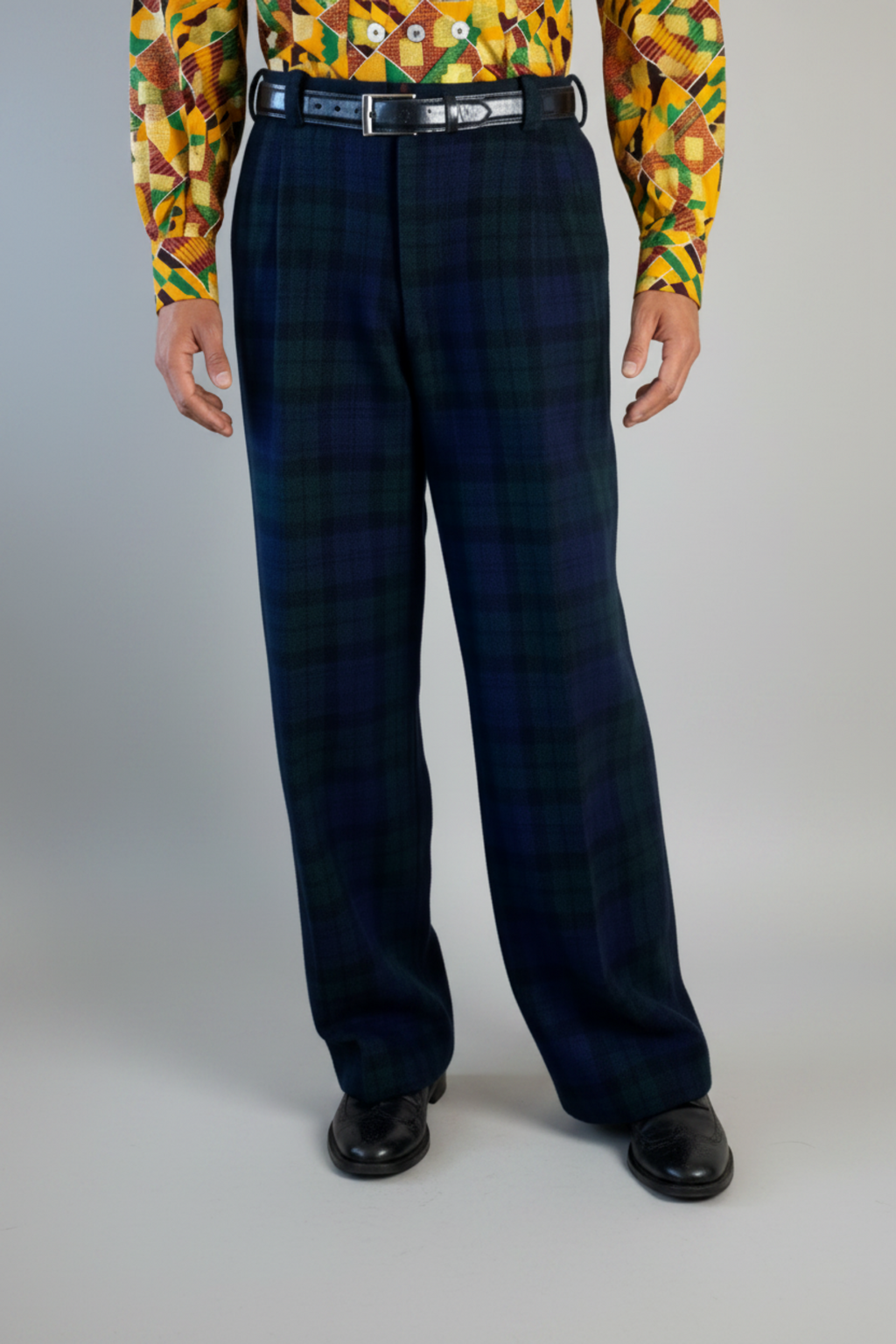 Stacy Stewart Smith's men's  Harris Tweed© Pleated Windsor Pants on a mannequin 6'#" cropped view front. Worn wiith the Noble Kente Kente cloth long-sleeve button-up shirt with a dense multicolor abstract print in yellow, orange, green, and red, with black shoes, standing against a gray backdrop.