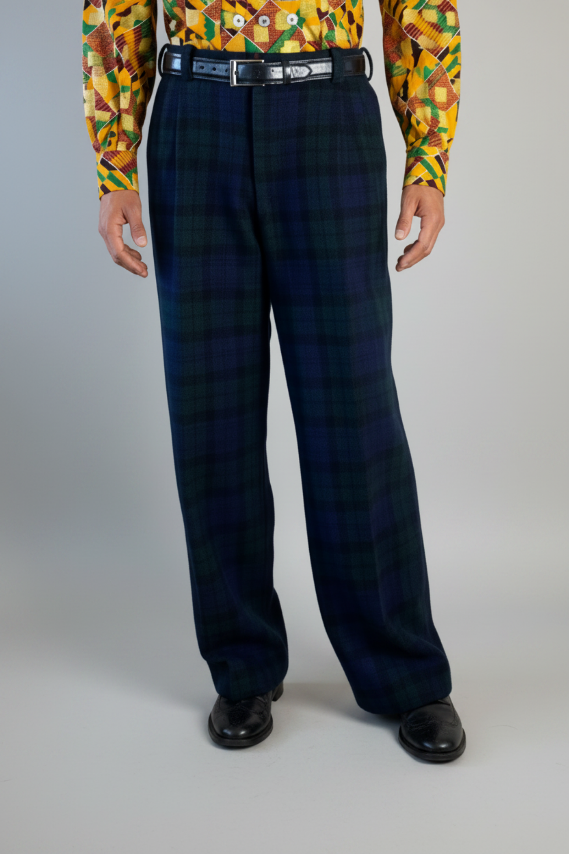 Stacy Stewart Smith's men's  Harris Tweed© Pleated Windsor Pants on a mannequin 6'#" cropped view front. Worn wiith the Noble Kente Kente cloth long-sleeve button-up shirt with a dense multicolor abstract print in yellow, orange, green, and red, with black shoes, standing against a gray backdrop.