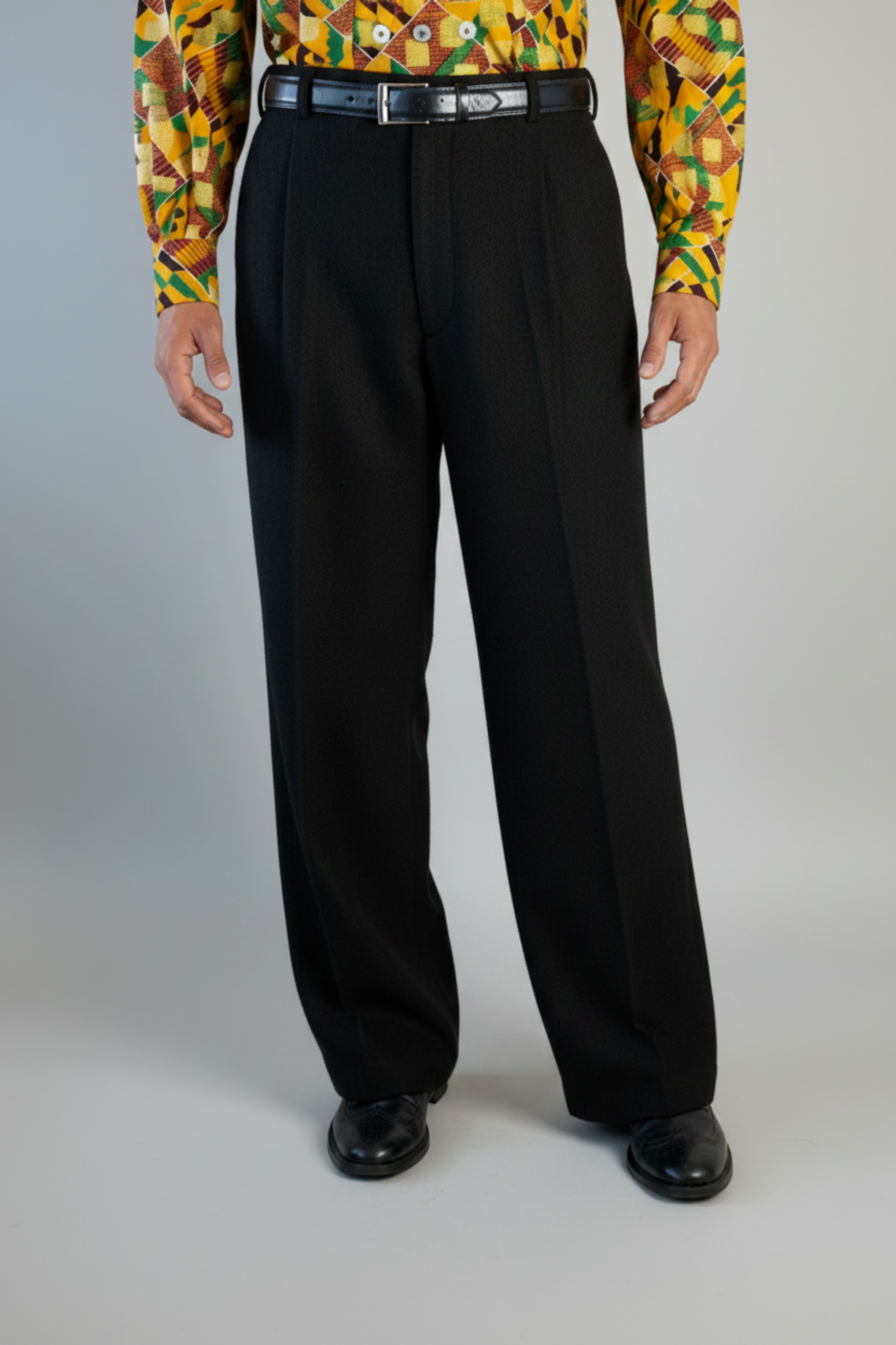 Stacy Stewart Smith's men's solid onyx Harris Tweed© Pleated Windsor Pants on a mannequin 6'3" cropped front view. Worn wiith the Noble Kente Kente cloth long-sleeve button-up shirt with a dense multicolor abstract print in yellow, orange, green, and red, with black shoes, standing against a gray backdrop.
