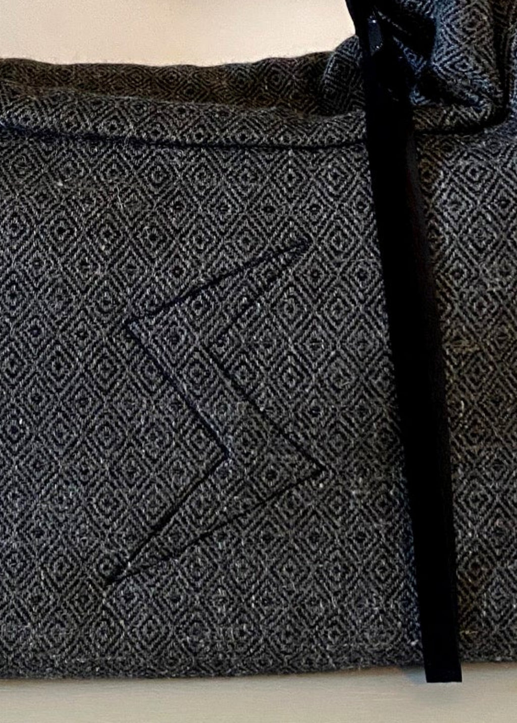 Stacy Stewart Smith's Men's Diamond Dodger Trapezoid messenger bag in Cement/Onyx zoomed frontal view of logo quilted embroidery against a gray background.