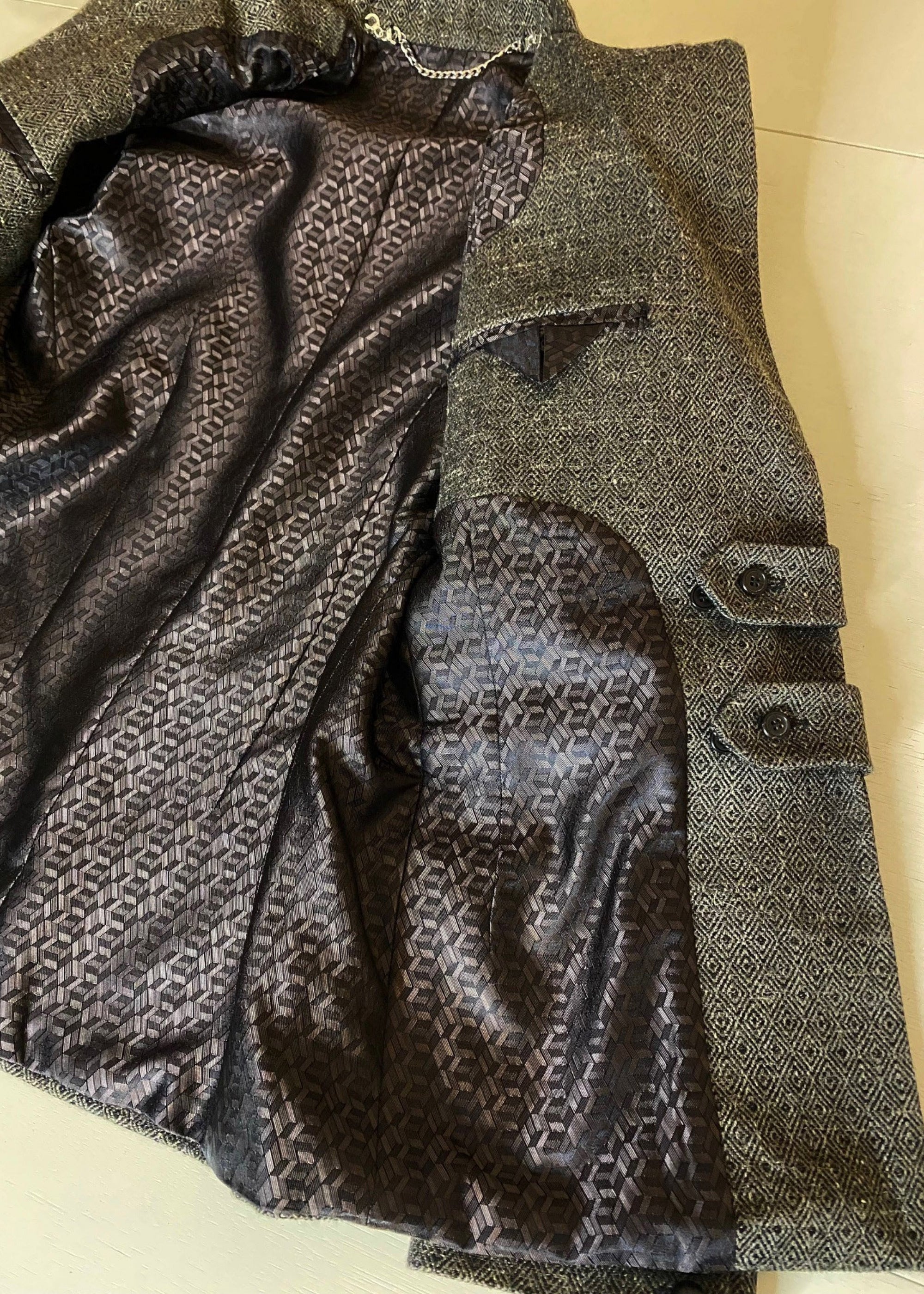 Stacy Stewart Smith Men's Highland Gentry Diamond Tweed Suit lying flat with one side of the front open revealing the box pattered acetate liningl and chain hanging accessory on an Ivory shadowed paper surface. 