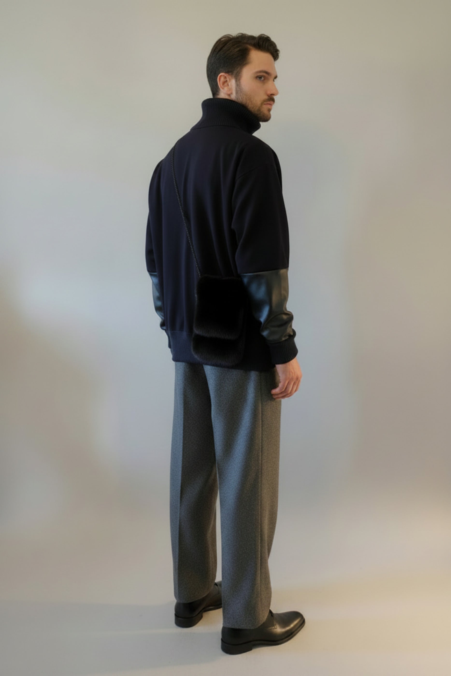 Handsome male model wearing the Harlequin Patch Sweatshirt and Diamond Dodger Pleated Tweed Windsor Pants has the designer's Mink Sporran bag over one shoulder from the back view against a light gray backdrop.