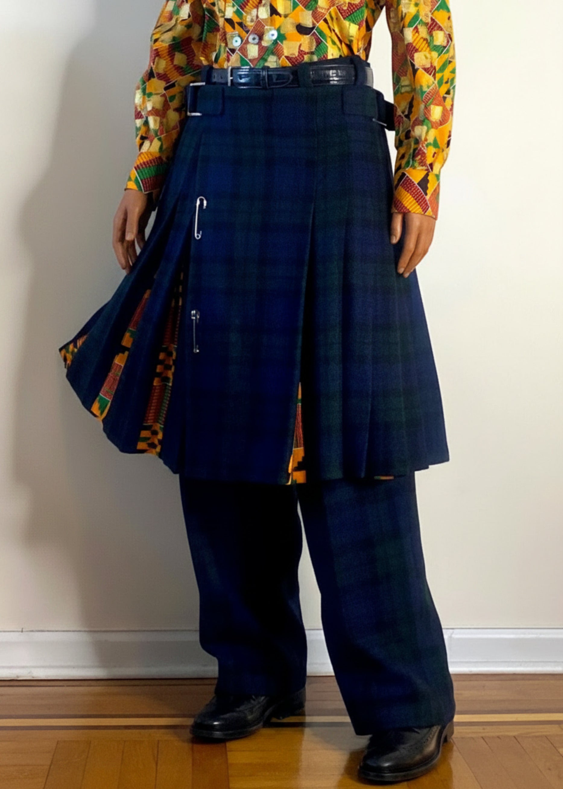 Stacy Stewart Smith's men's  Harris Tweed© Pleated Windsor Pants on a mannequin 6'3" front view. Worn with the Noble Kente Kente cloth long-sleeve button-up shirt with a dense multicolor abstract print in yellow, orange, green, and red, with the Blackwatch Plaid Harris Tweed© Reality Kilt flowing to show kente cloth pattern inlay with black shoes, standing against a gray backdrop.