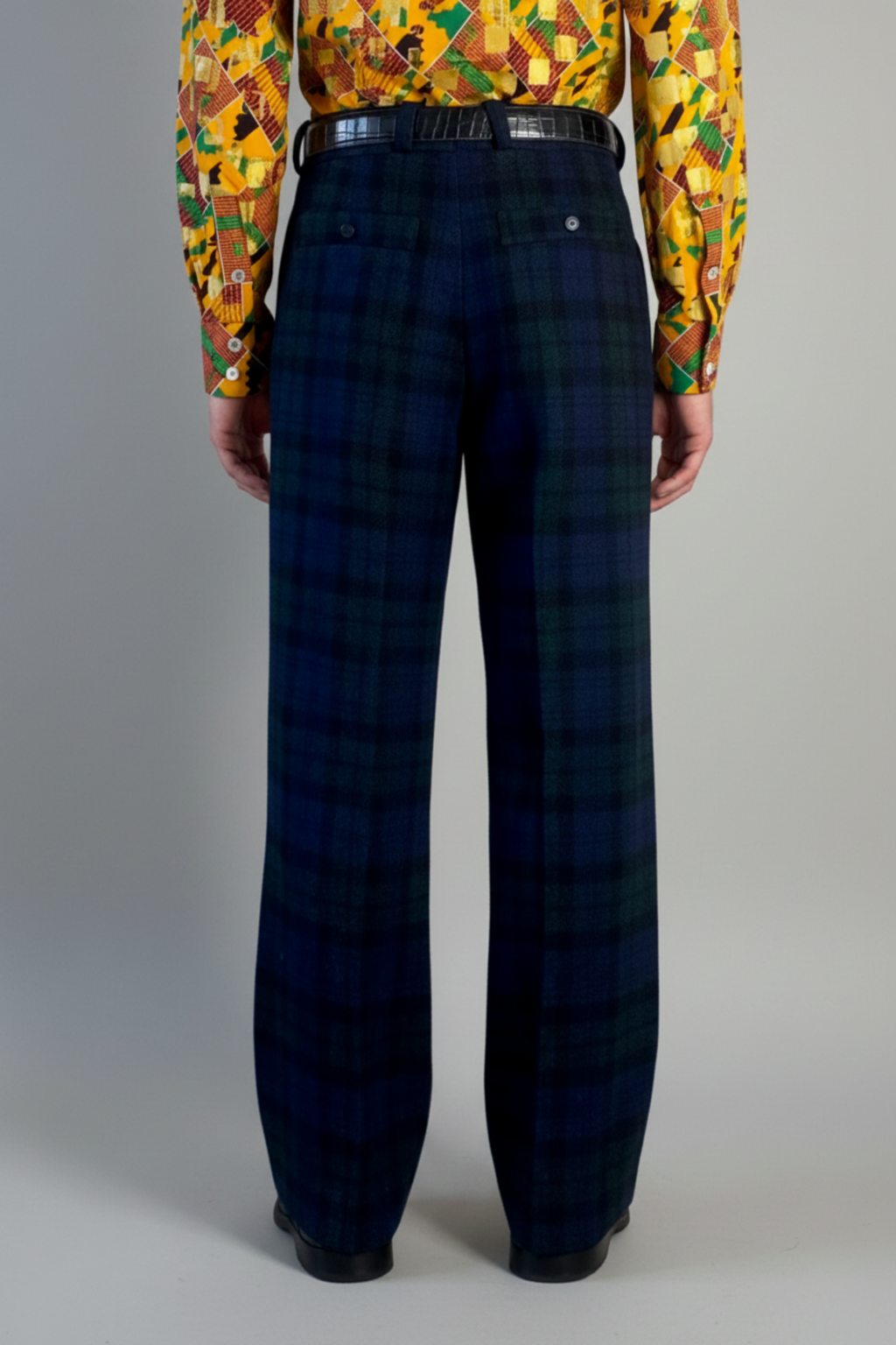 Stacy Stewart Smith's men's  Harris Tweed© Pleated Windsor Pants on a mannequin 6'3" cropped back view. Worn with the Noble Kente cloth long-sleeve button-up shirt with a dense multicolor abstract print in yellow, orange, green, and red, with black shoes, standing against a gray backdrop.