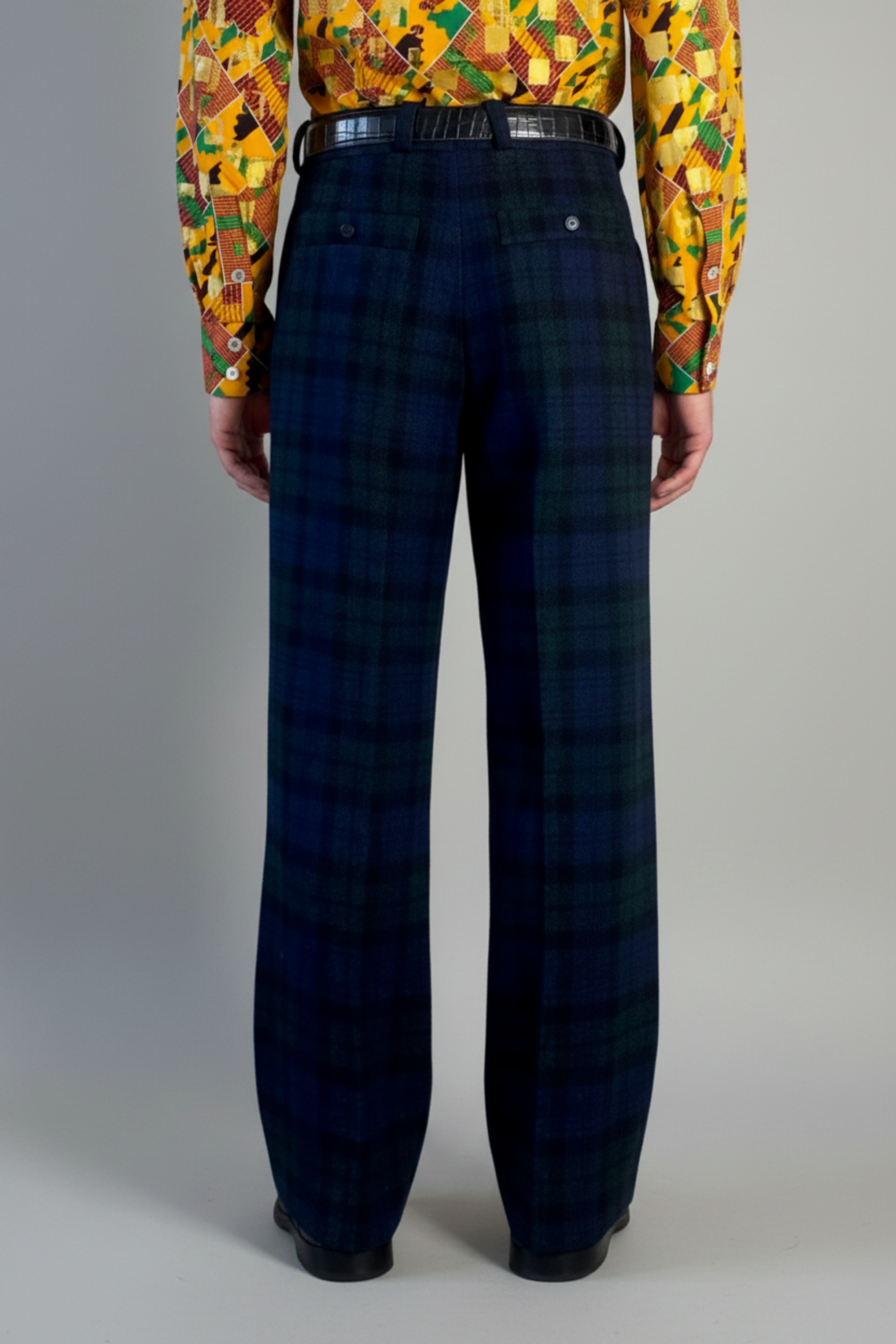 Stacy Stewart Smith's men's  Harris Tweed© Pleated Windsor Pants on a mannequin 6'3" cropped back view. Worn with the Noble Kente cloth long-sleeve button-up shirt with a dense multicolor abstract print in yellow, orange, green, and red, with black shoes, standing against a gray backdrop.
