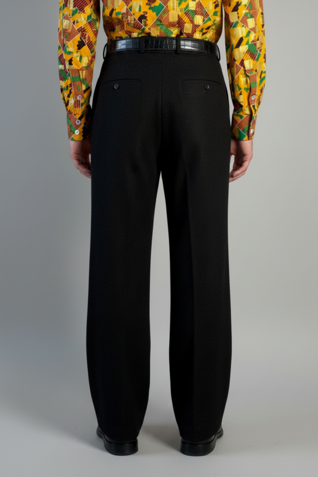 Stacy Stewart Smith's men's solid onyx  Harris Tweed® Pleated Windsor Pants on a mannequin 6'3" cropped back view. Worn with the Noble Kente cloth long-sleeve button-up shirt with a dense multi-color abstract print in yellow, orange, green, and red, with black shoes, standing against a gray backdrop.