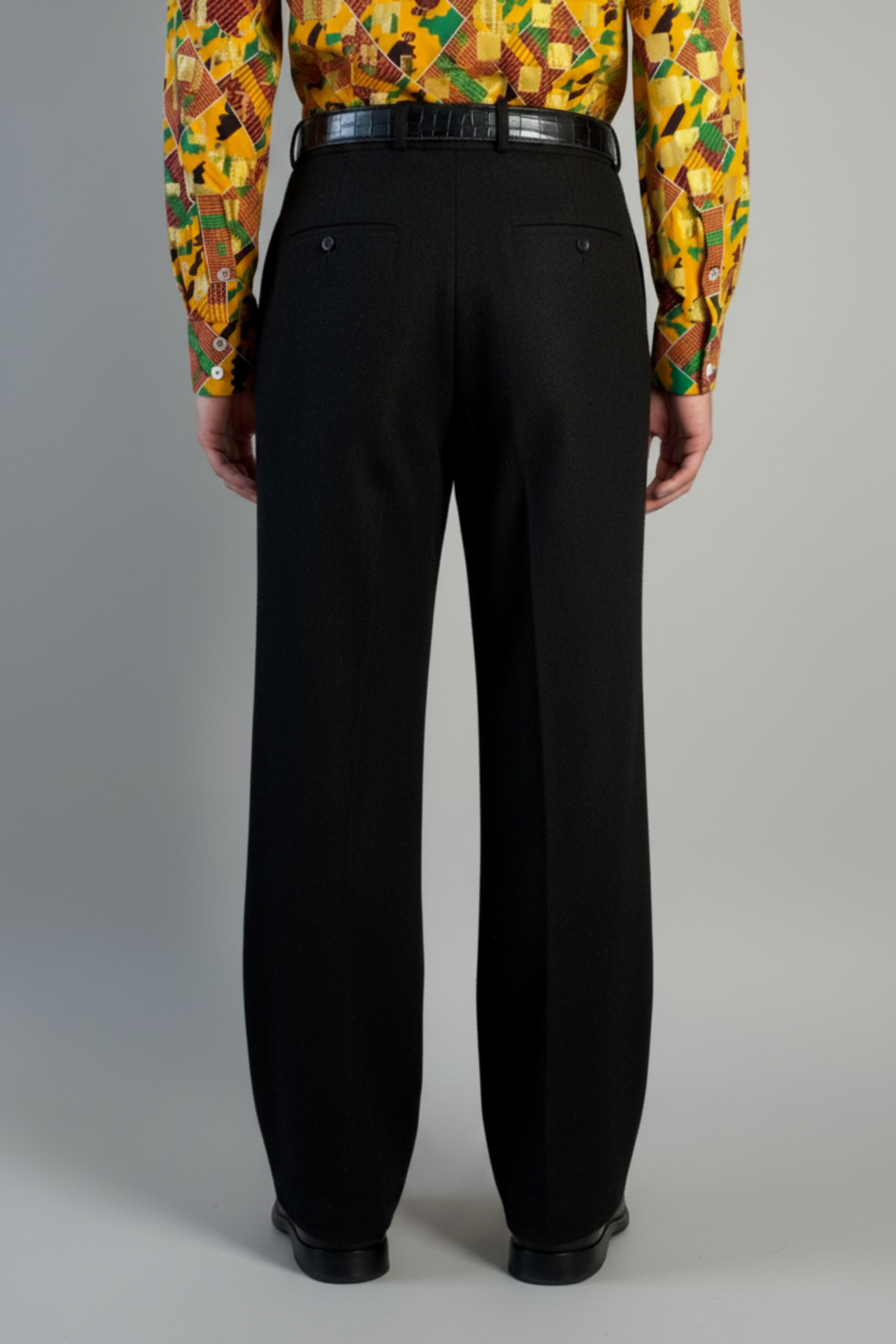 Stacy Stewart Smith's men's solid onyx  Harris Tweed© Pleated Windsor Pants on a mannequin 6'3" cropped back view. Worn with the Noble Kente cloth long-sleeve button-up shirt with a dense multicolor abstract print in yellow, orange, green, and red, with black shoes, standing against a gray backdrop.