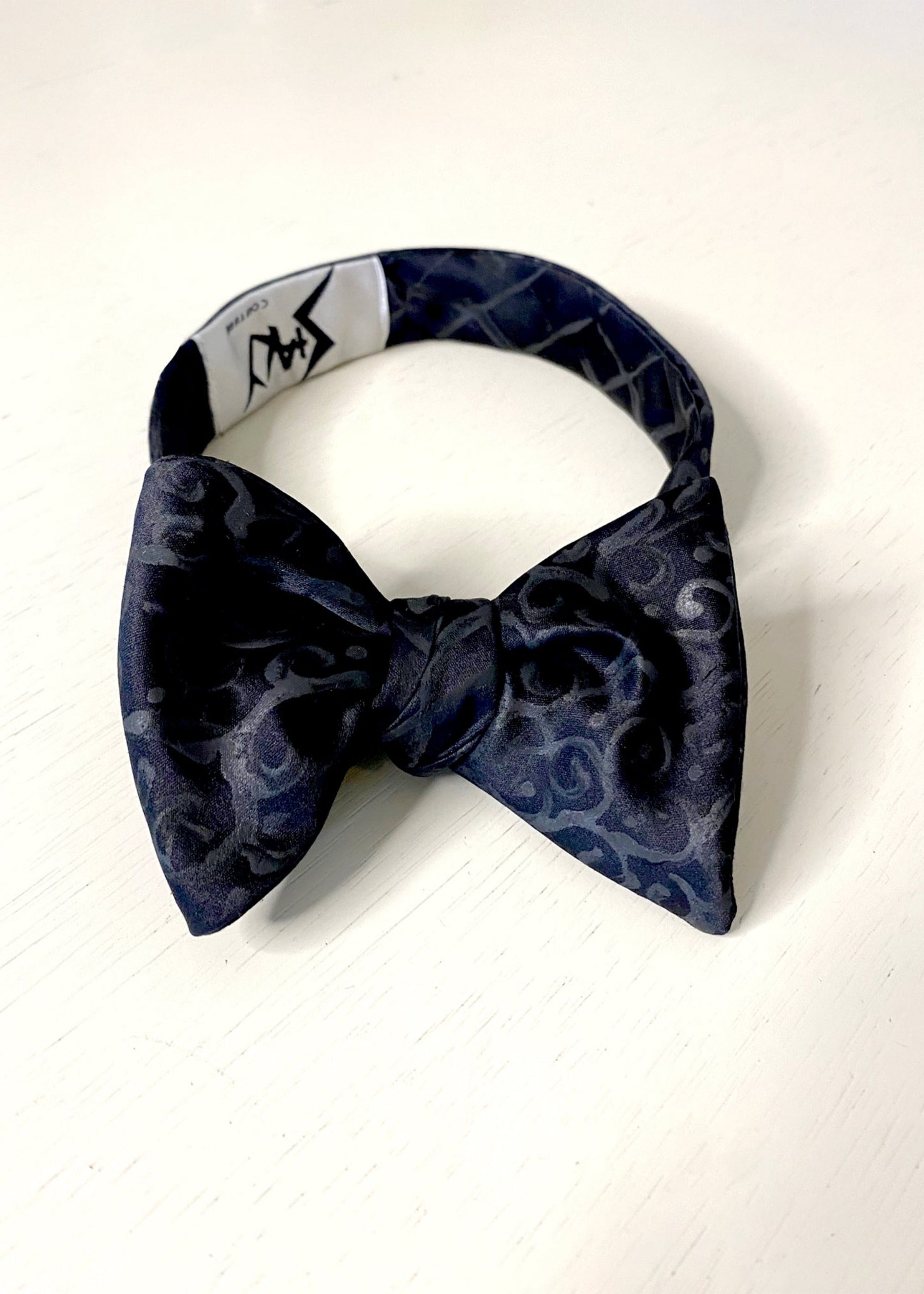 A silk hand-painted black arabesque patterned bow tie by Stacy Stewart Smith, front view showing the brand label against a neutral background.