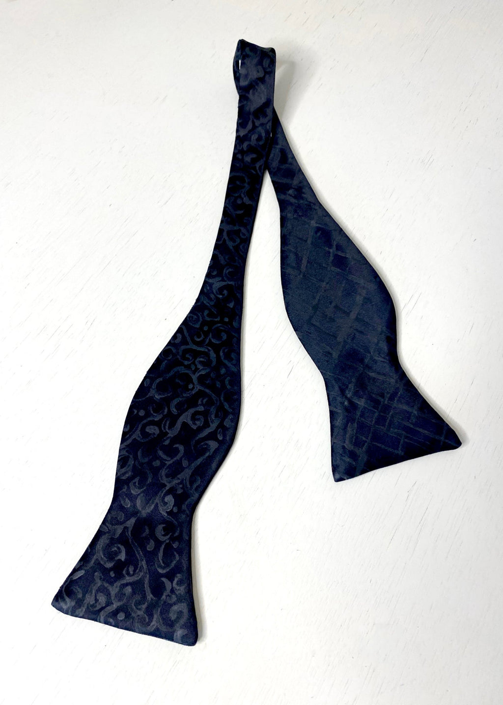 A silk hand-painted black arabesque and plaid patterned bow tie by Stacy Stewart Smith, untied view against a neutral background.