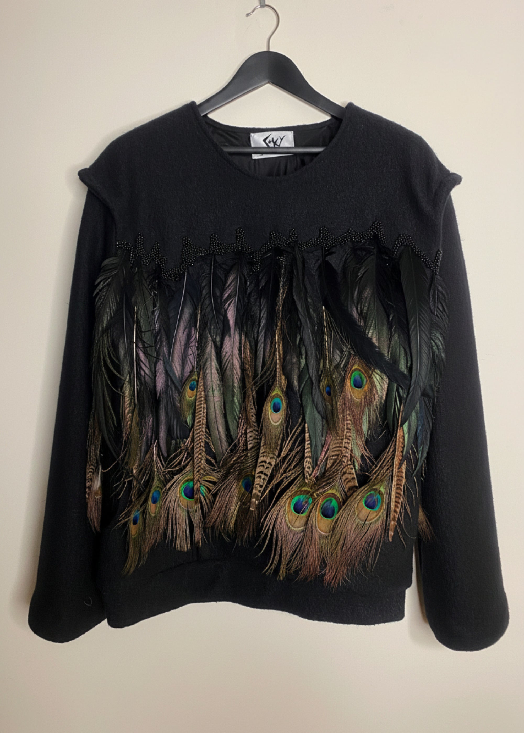 Stacy Stewart Smith Men's Pennaceous Beaded Black Boiled Wool Sweater with peacock, quail and rooster feathers on a hanger front view  against an ivory backdrop. 
