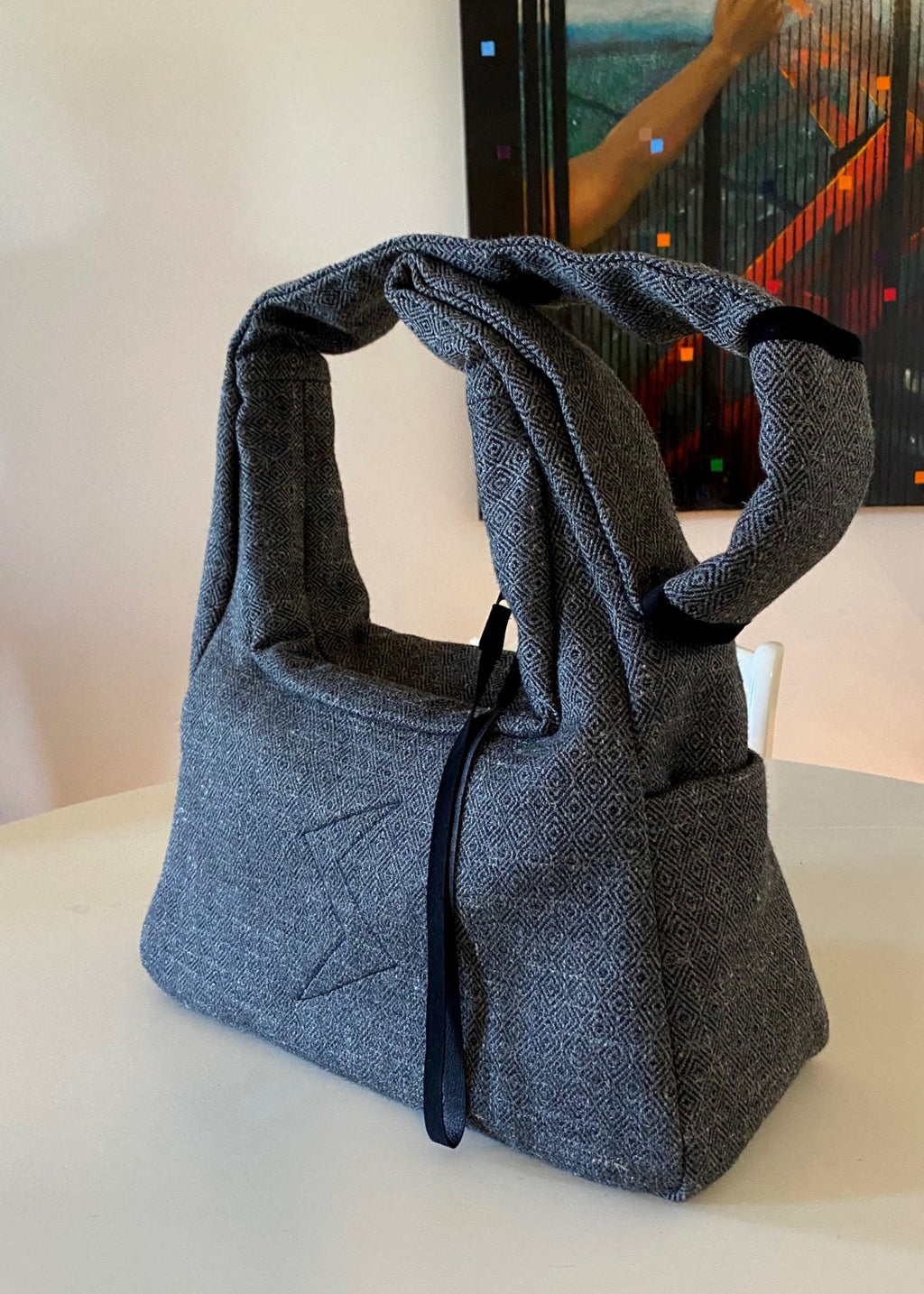 Stacy Stewart Smith's Men's Diamond Dodger Trapezoid messenger bag in Cement/Onyx 3/4 view on an ivory table in a room with an ivory wall with an oil painting by the designer hanging on it.