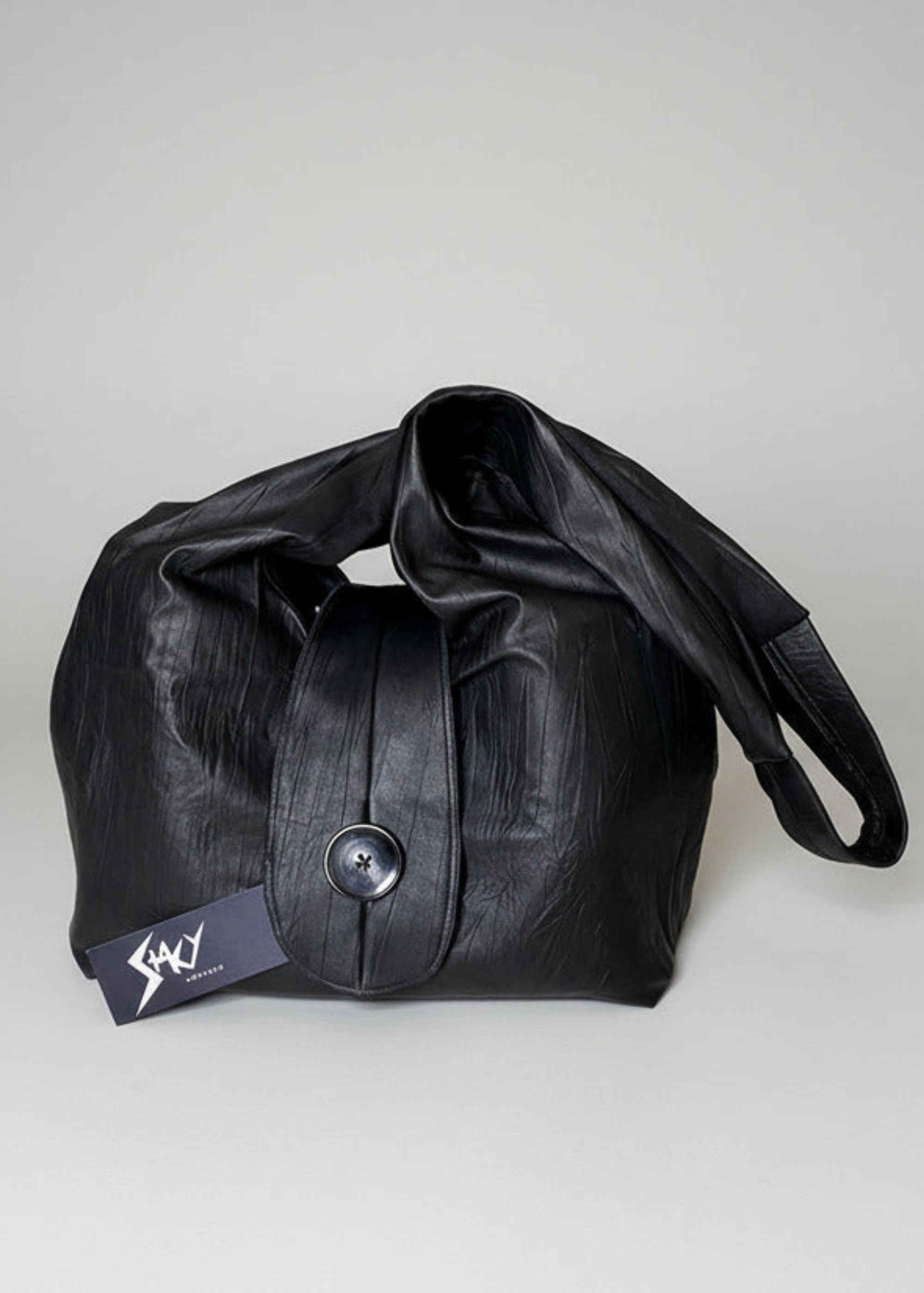 Stacy Stewart Smith Euclidian Space Crinkled Leather Messenger Bag frontal view frontal view with tongue flap and brand hang tag in front of a gray backdrop.