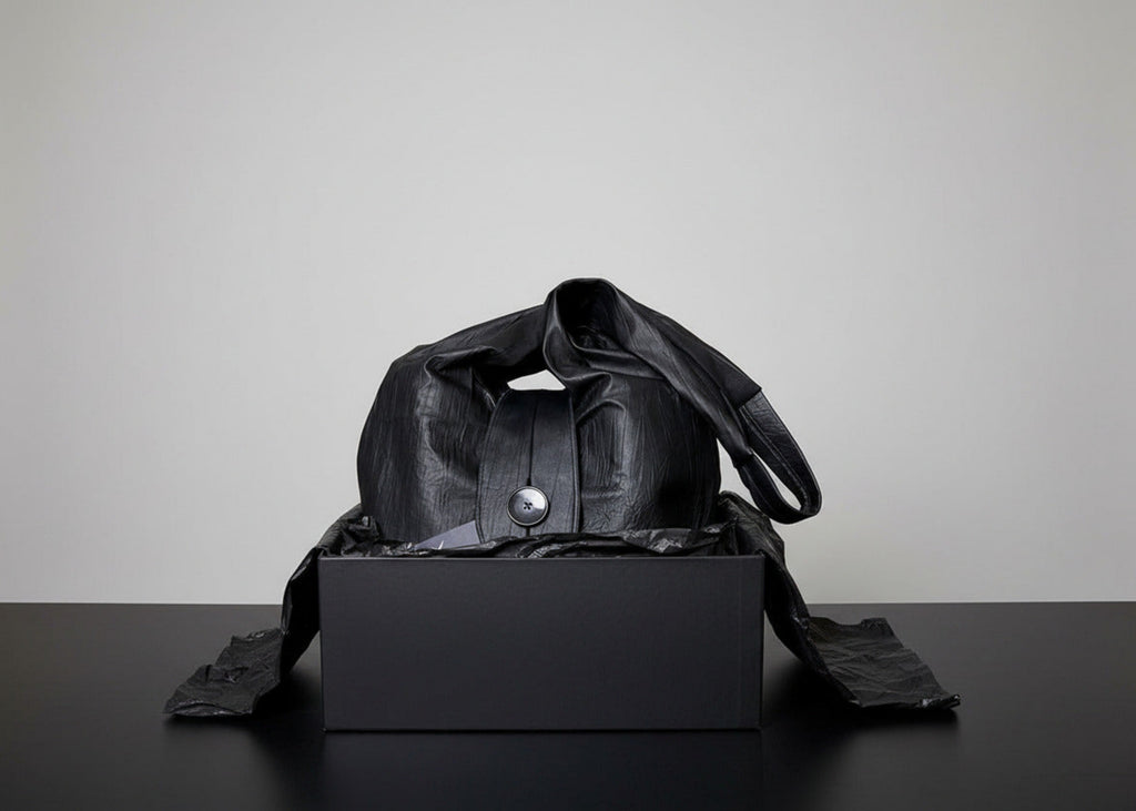 Stacy Stewart Smith Euclidian Space Crinkled Leather Messenger Bag frontal view in a black gift box with tongue flap showing and self-handle wrapped over the top and black tissue paper around it on a black surface with a gray backdrop. 