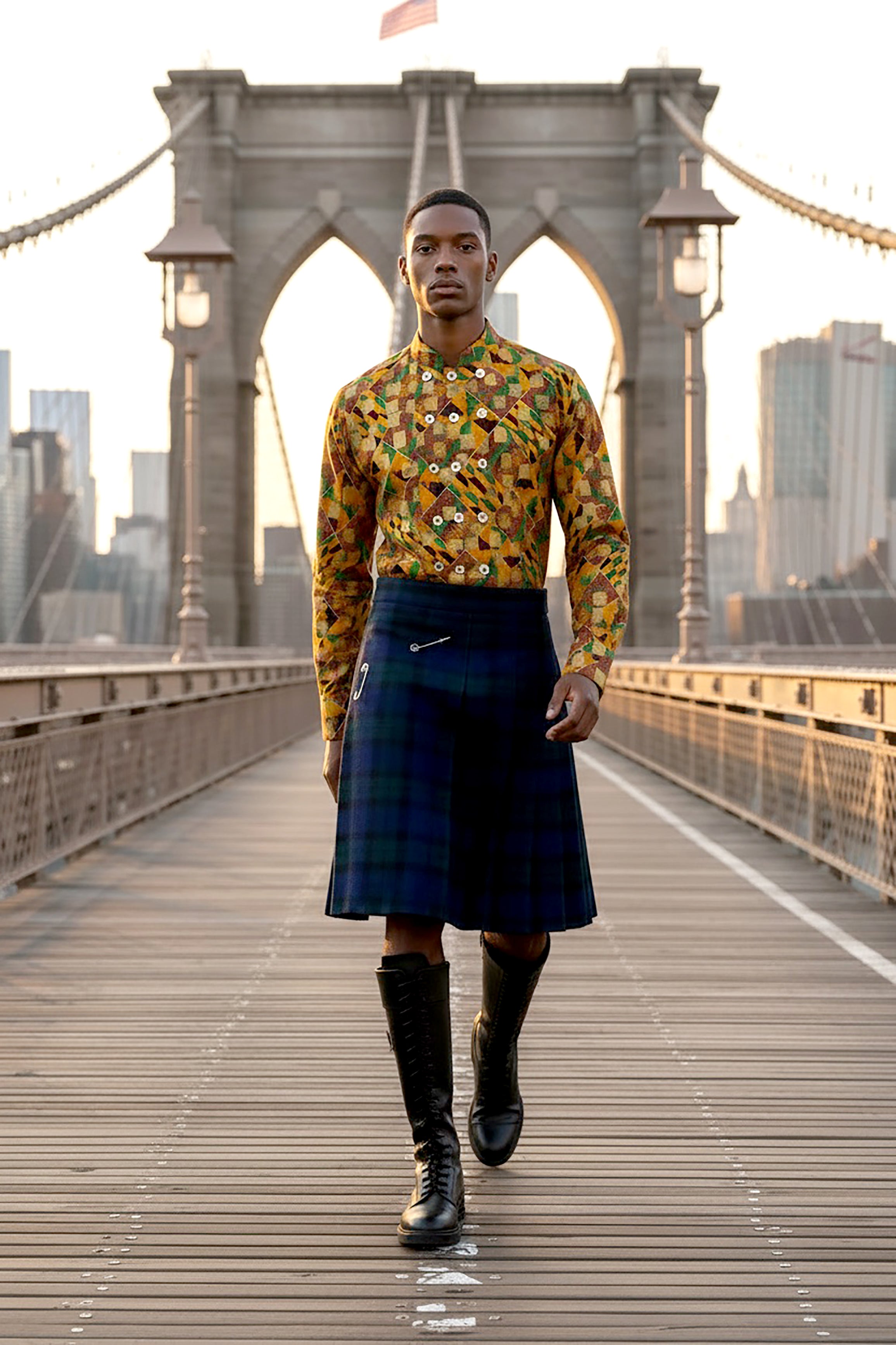Stacy Stewart Smith's Noble Kente long-sleeve button-up premium cotton Kente cloth dress shirt with manadarin collar, long sleeve gold metallic hand pixel over-printing and real shell buttons styled with Blackwatch plaid Reality Kilt on a male model walking on the Brooklyn Bridge.