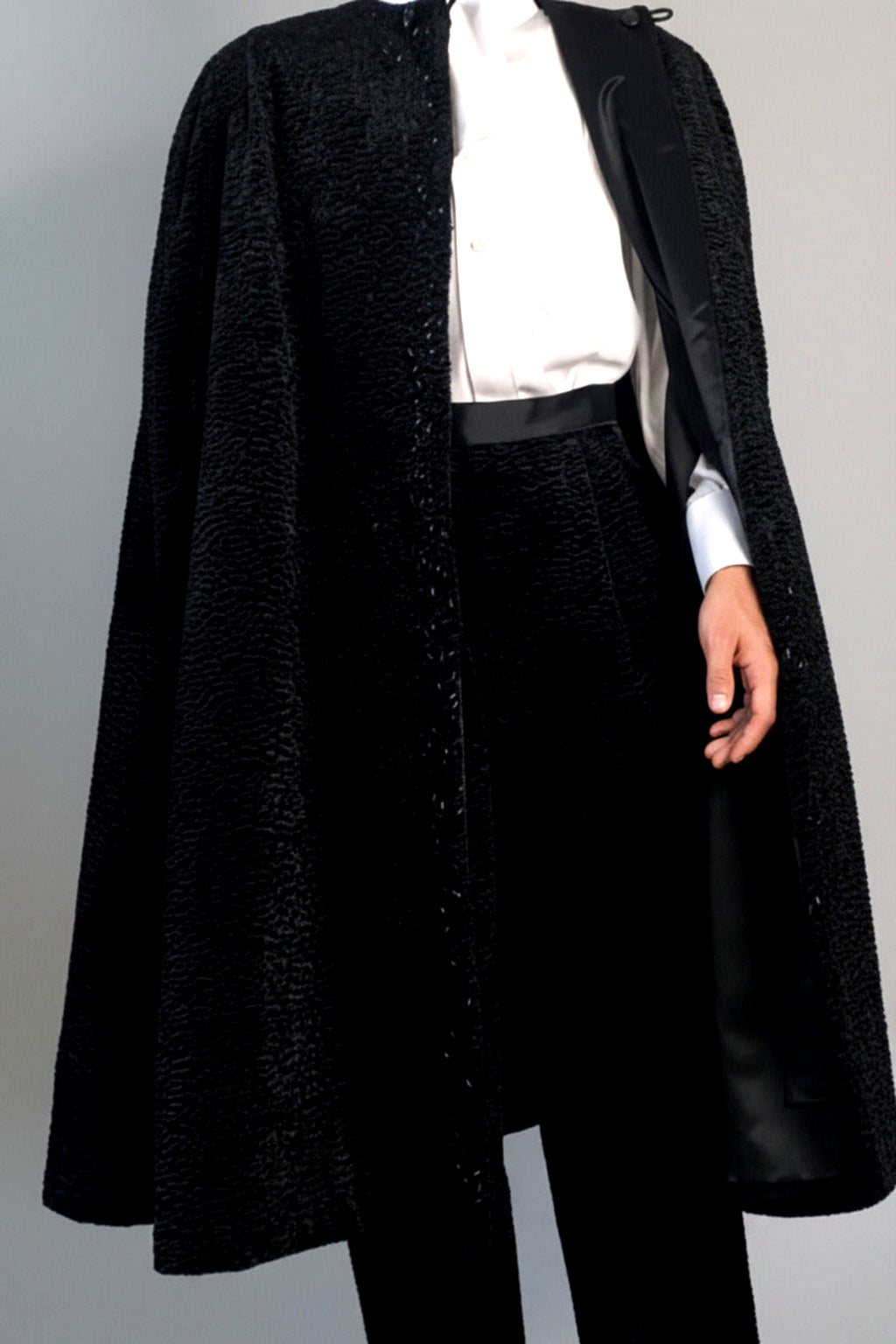 The Grand Soir Palace Cape by Stacy  Stewart Smith is cut for exactly that moment — a structured sweep of flowing black Persian lamb on a mannequin 6'3" against a gray backdrop. Front view with  cape fully open to show bugle beading, neckline loops, and one arm through inside satin lining and holder, which also reveals the matching Grand Soir Tuxedo trousers.