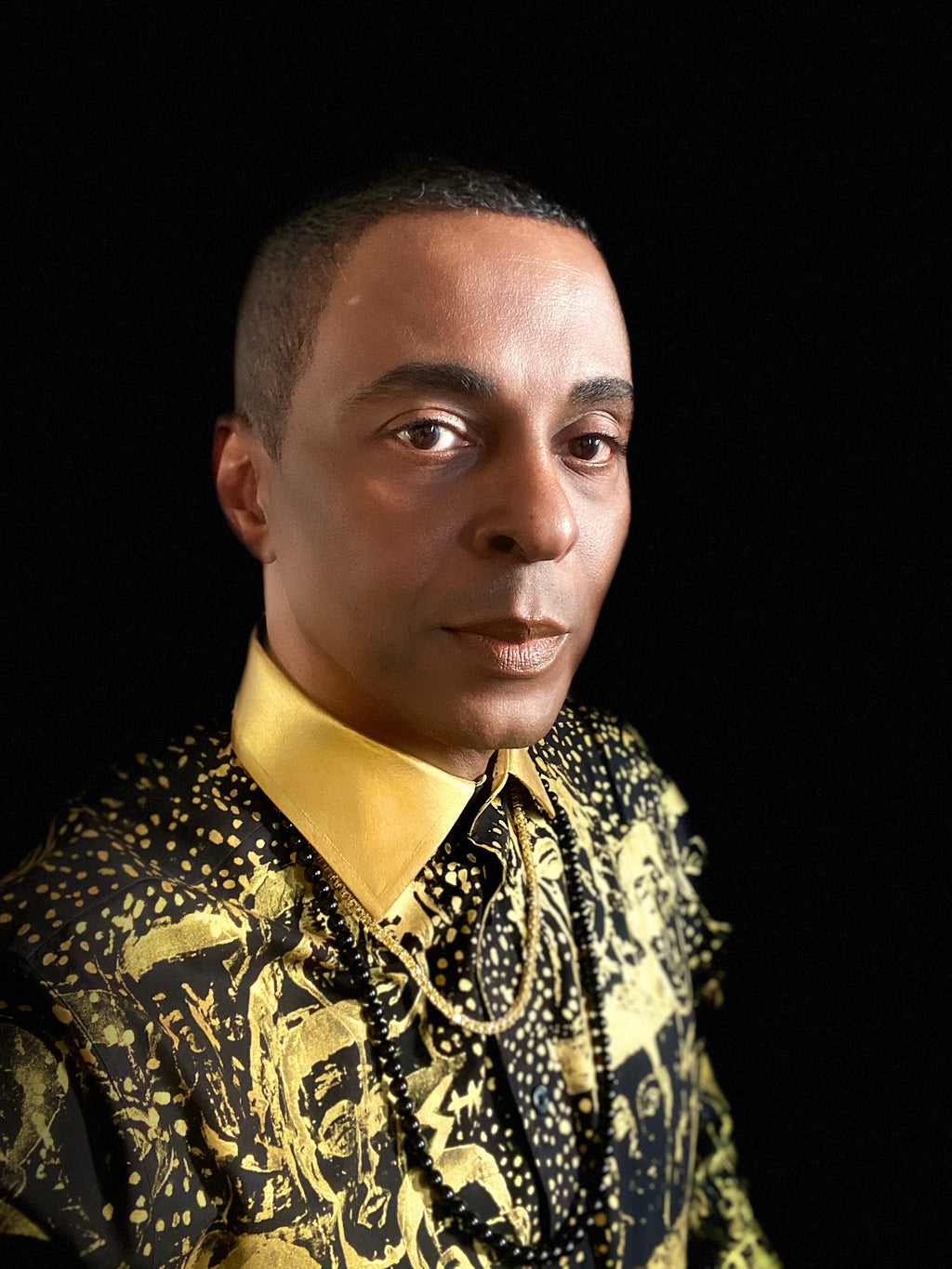 A close-up portrait of couturier, fine artist and author, Stacy Stewart Smith wearing  his Paws in Gold hand-painted and printed black premium cotton percale shirt with a gold metallic collar, set against a solid black background.