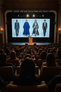 A large auditorium filled with seated audience members facing a stage where a presenter stands behind a podium in front of a giant screen showing four fashion models in formal and long coats.
