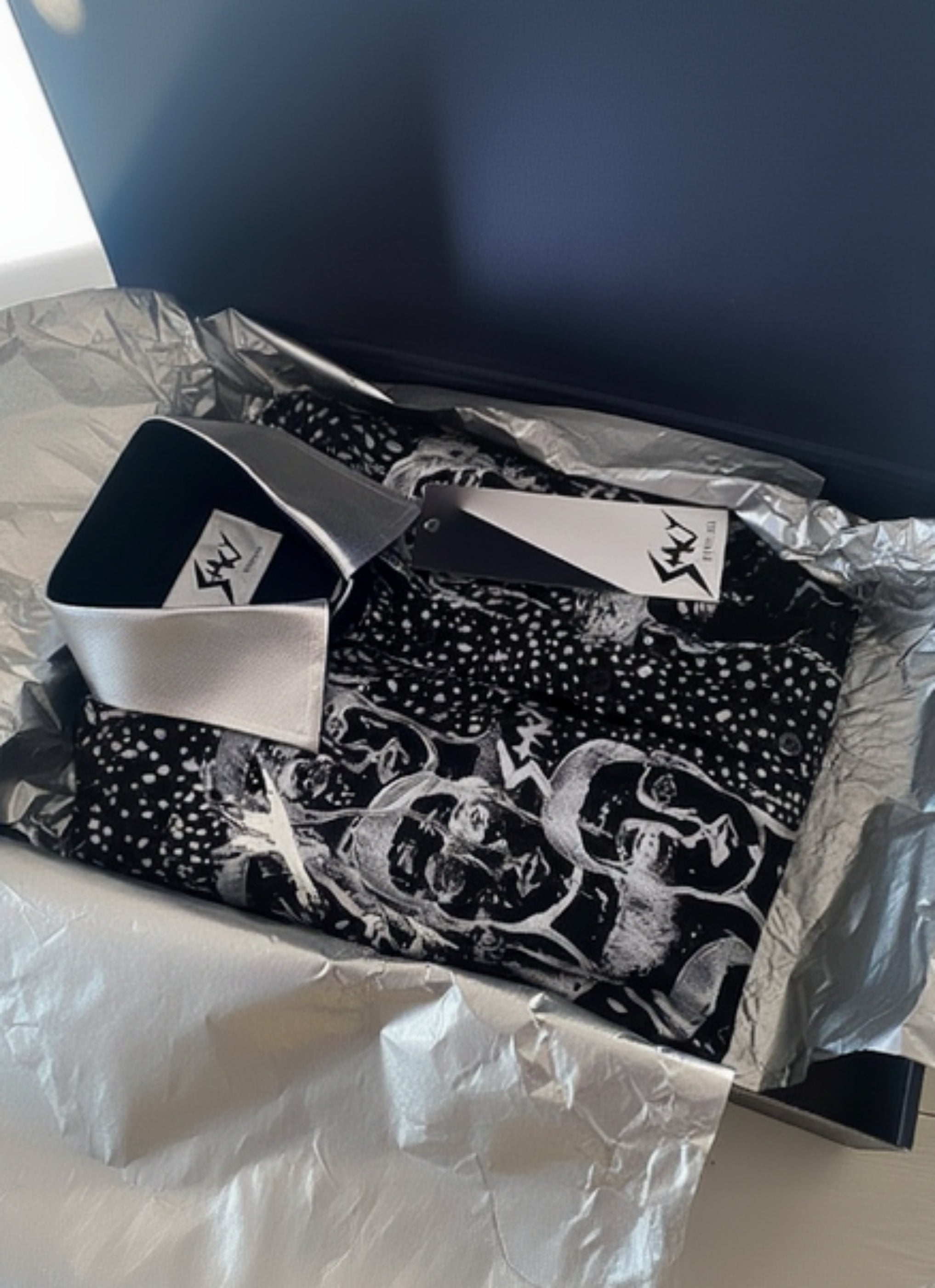 Stacy Stewart Smith Men’s Pawns in Silver on Black Hand-Painted Premium Cotton Percale Dress Shirt folded in a black giift box with gold tissue paper and brand label and tag showing on a light colored surface. 