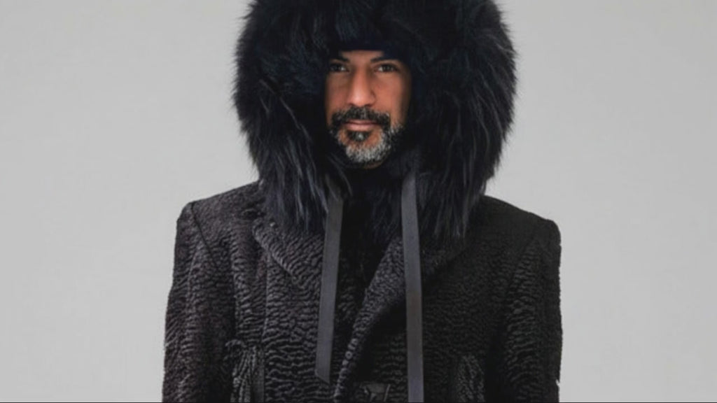 Front view of a man wearing the black Grand Soir Expedition Coat by Stacy Stewart Smith, with the hood’s fur trim and ribbon ties moving gently.